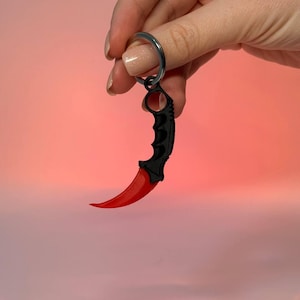 May include: A black and red karambit knife keyring. The keyring features a curved red blade and a black handle with finger grooves. The keyring is attached to a silver key ring. The background is a gradient of pink and orange.