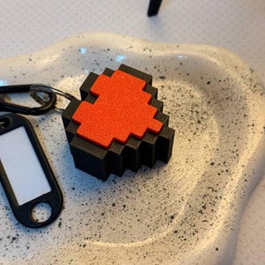 Peut inclure: Un porte-clés en forme de cœur pixelisé noir et rouge. Le porte-clés comporte une petite étiquette rectangulaire avec un espace blanc. Le cœur est une conception imprimée en 3D, et le porte-clés est attaché à un anneau argenté.