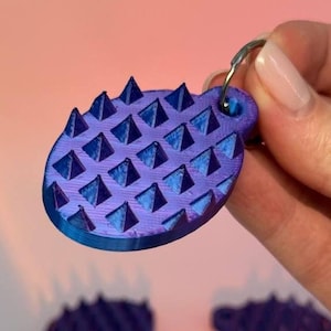 Op de afbeelding: Een paarse en blauwe sleutelhanger met een textuur van verhoogde piramidevormen. De sleutelhanger wordt vastgehouden door een zilveren ring, een uniek accessoire.