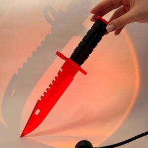 Pode incluir: Uma faca vermelha e preta com uma lâmina serrilhada e um cabo preto. A faca tem uma guarda e uma ponta vermelha. A faca é segurada por uma mão. A faca é iluminada por uma luz vermelha.