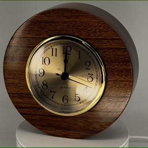 May include: A round, wooden quartz clock with a gold-colored face and hands. The clock face features black numerals and the word "QUARTZ" at the bottom. The clock is encased in a dark brown, circular wooden frame.