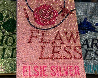 以下が含まれることがあります： Elsie Silverの「Flawless」というタイトルのピンク色の本。キラキラ光るラインストーンで装飾されています。表紙にはバラと帽子のデザインがあります。本は金色のスタンドに展示されており、背景にはラインストーンで覆われた他の本があります。