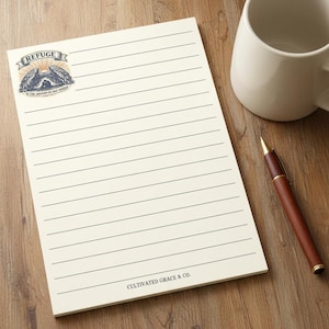 May include: A cream-colored notepad with horizontal black lines. The top left corner features a graphic of wings and the text REFUGE. The bottom center displays the text CULTIVATED GRACE & CO. A brown and gold pen and a white mug are nearby.