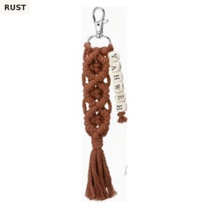May include: A rust-colored macrame keychain featuring a silver metal lobster clasp, a knotted cord design, and a dangling strand of white square wooden beads with black letters spelling YAHWEH, finished with a matching rust-colored cord tassel.
