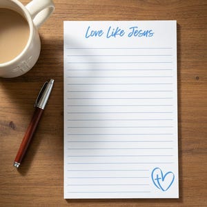 Può includere: Un blocco note rettangolare bianco con linee blu orizzontali, caratterizzato dal testo blu Love Like Jesus nella parte superiore e da un'icona a forma di cuore blu con una croce all'interno nell'angolo in basso a destra, con una penna argentata e color legno.