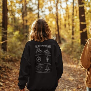 Op de afbeelding: Zwart sweatshirt met de tekst "THE WEEKEND FORECAST" en illustraties van wandelschoenen, bergen, een kompas, een boom, een snackdrager en een kampvuur. Het sweatshirt wordt buiten gedragen.