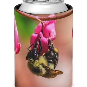 May include: A can cooler featuring a close-up image of a bee clinging to a pink flower. The bee is black and yellow, and the flower is a vibrant pink. The background is a soft, blurred pink and green.