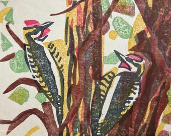 May include: A colorful woodblock print featuring two woodpeckers perched on tree branches. The birds have red crests, black and white bodies, and are surrounded by green and yellow foliage. The artwork has a rustic, handmade aesthetic.