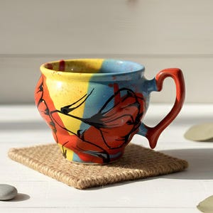 Könnte beinhalten: Eine farbenfrohe Keramiktasse mit einem einzigartigen Design. Die Tasse hat eine lebendige Mischung aus Gelb, Blau und Rot, akzentuiert mit schwarzen Linien. Sie steht auf einem quadratischen, gewebten Untersetzer. Der Griff ist leuchtend rot.