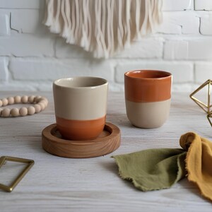 Pode incluir: Canecas de cerâmica de duas cores em bege e laranja, uma em uma base de madeira. Uma pulseira de contas de madeira, uma forma geométrica dourada e tecido também estão sobre a mesa. O fundo apresenta uma parede de tijolos brancos e uma tapeçaria tecida.