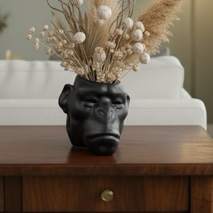Puede incluir: Un jarrón de cerámica negra con forma de cabeza de gorila, lleno de flores y hierbas secas. El jarrón está sobre una mesa de madera oscura. El arreglo incluye hierba de la pampa beige y acentos esféricos blancos.