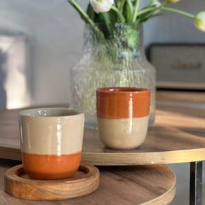 May include: Two-tone ceramic mugs in beige and orange, one on a wooden coaster, sit on a round wooden table. A clear glass vase with white tulips is in the background. The table has black metal legs. The scene evokes a sense of calm.