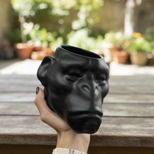 Puede incluir: Una maceta de cerámica negra con forma de cabeza de chimpancé. La maceta tiene un acabado liso y mate. La imagen muestra la maceta sostenida en una mano. El fondo está borroso, mostrando una mesa de madera y vegetación.