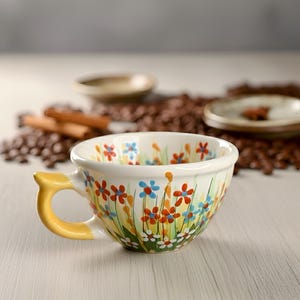 Può includere: Una tazza da tè in ceramica bianca con manico giallo, decorata con un motivo floreale di fiori rossi, blu e arancioni. La tazza è circondata da chicchi di caffè e piattini.