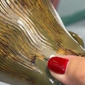 Puede incluir: Primer plano de una taza de cer&aacute;mica con un patr&oacute;n texturizado en forma de olas en tonos marr&oacute;n, dorado y verde. La superficie de la taza tiene un acabado brillante. Una mano con esmalte de u&ntilde;as rojo toca la taza.