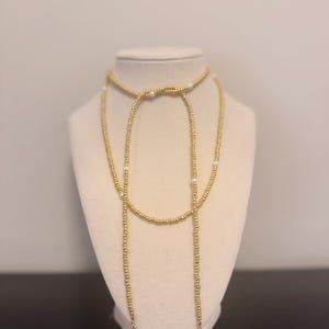 May include: A gold beaded lariat necklace with white pearl accents and a pearl drop. The necklace is displayed on a mannequin. The gold beads are strung together to create a long, elegant design.