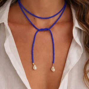 May include: A long, thin necklace made of small, vibrant blue beads. The necklace wraps around the neck once and features a central knot, with two long strands hanging down, each finished with a single, irregular white pearl at the end.