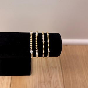 Puede incluir: Cuatro pulseras doradas con detalles blancos se exhiben en un soporte de terciopelo negro. Las pulseras varían en diseño, algunas con pequeñas cuentas y otras con elementos blancos más prominentes.