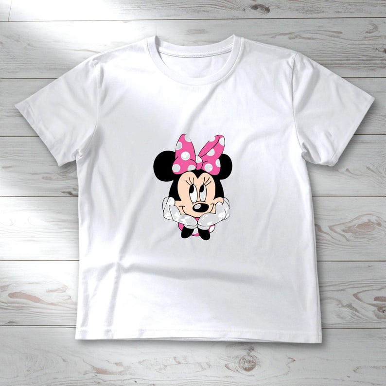 May include: White t-shirt featuring a graphic of Minnie Mouse. Minnie Mouse is depicted with black ears, a pink polka-dot bow, and a smiling face. The shirt is made of a soft, lightweight material, perfect for casual wear.
