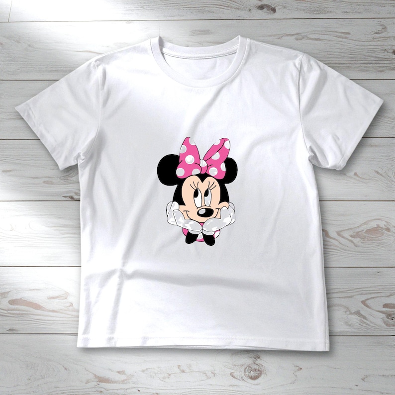 May include: White t-shirt featuring a graphic of Minnie Mouse. Minnie Mouse is depicted with black ears, a pink polka-dot bow, and a smiling face. The shirt is made of a soft, lightweight material.