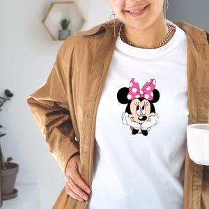 May include: White t-shirt featuring a Minnie Mouse graphic. The cartoon character has a pink polka dot bow and is smiling. The shirt is worn with a brown jacket and gold jewelry.