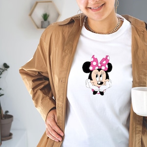 May include: White t-shirt featuring a Minnie Mouse graphic. The cartoon character has a pink polka-dot bow and is resting her chin on her hands. The shirt is worn with a brown jacket and gold jewelry.