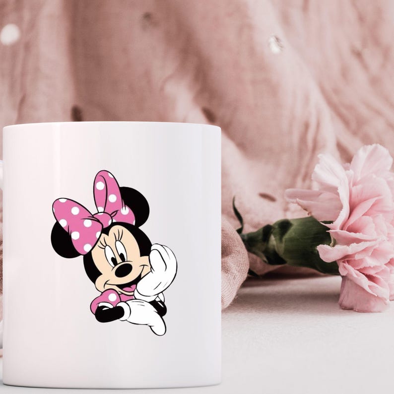 May include: White ceramic mug featuring a Minnie Mouse graphic. The cartoon character is depicted with a pink polka-dot bow, dress, and accents. A pink flower and fabric are in the background, adding a touch of color to the design.