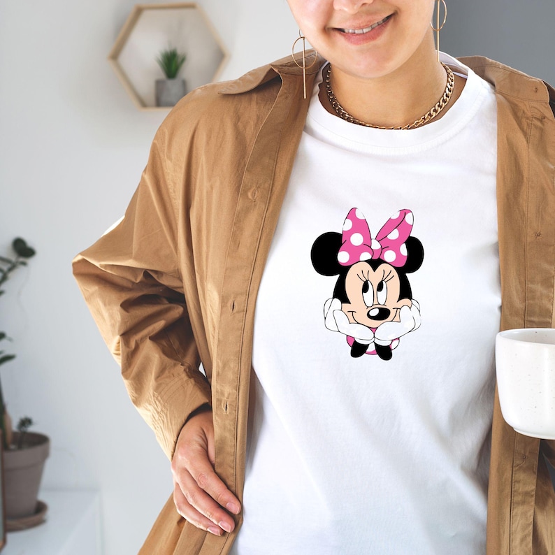 May include: White t-shirt featuring a Minnie Mouse graphic. The cartoon character has a pink bow with white polka dots and is smiling. The shirt is worn with a brown jacket and gold jewelry.