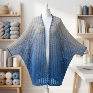 May include: A handmade, open-front cardigan in a gradient of gray to blue. The crochet design features a textured stitch and is trimmed with a darker blue border. The cardigan is displayed on a mannequin.