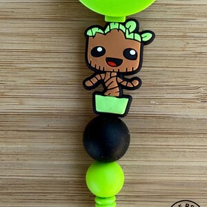 May include: A lime green retractable badge reel featuring a cartoon character shaped like a small brown tree in a green pot. The design includes a matte black bead and a lime green bead below the character, with a metal clip at the bottom.