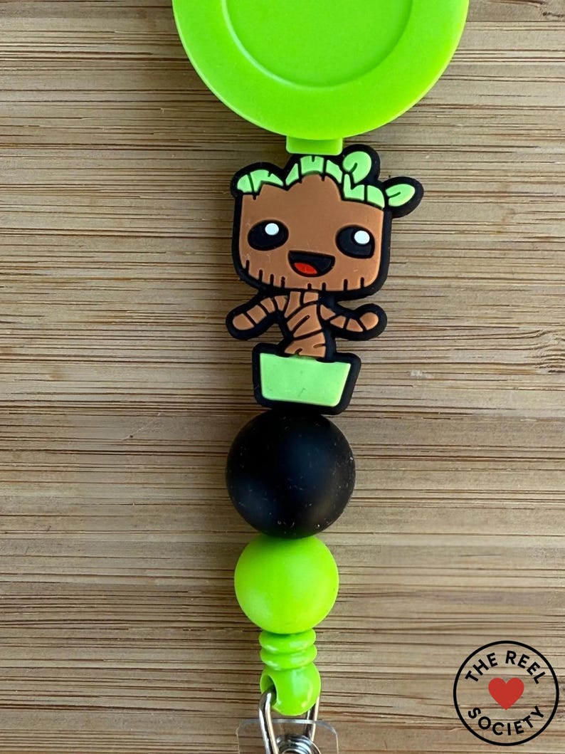 May include: A lime green retractable badge reel featuring a cartoon character shaped like a small brown tree in a green pot. The design includes a matte black bead and a lime green bead below the character, with a metal clip at the bottom.