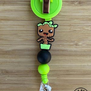 May include: A lime green retractable badge reel featuring a brown tree-like character figure with black eyes and a smile. The design includes a black bead and a lime green bead, finished with a clear plastic strap and a metal snap button closure.