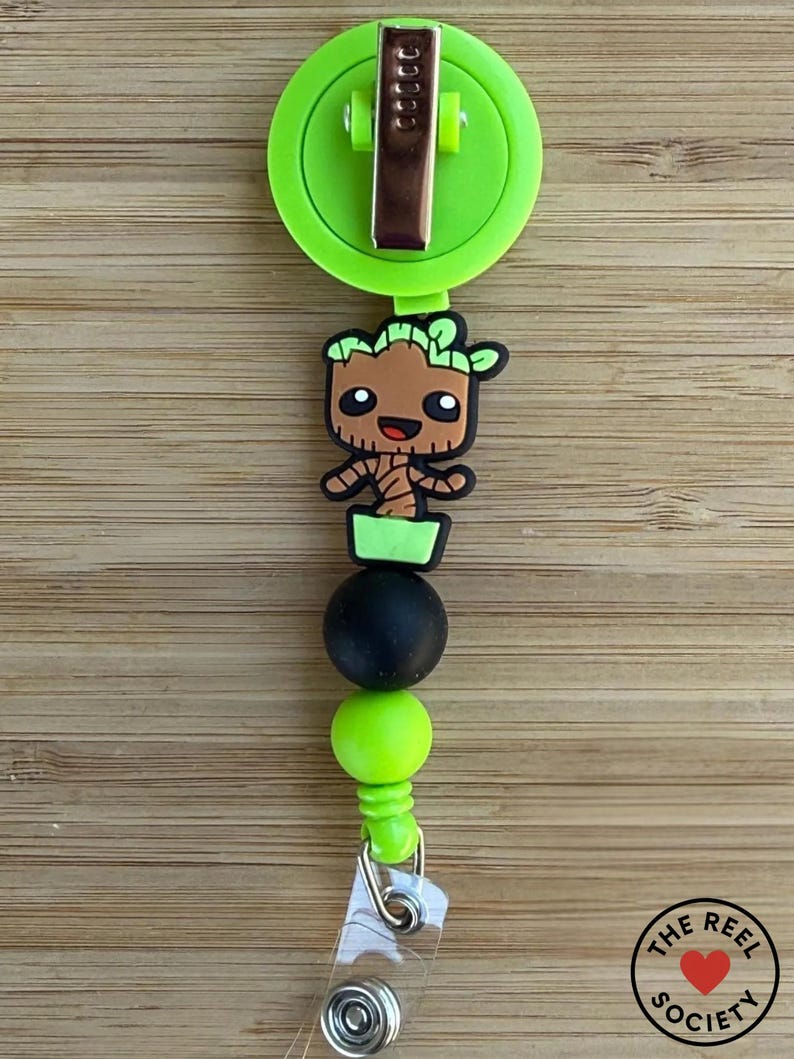 May include: A lime green retractable badge reel featuring a brown tree-like character figure with black eyes and a smile. The design includes a black bead and a lime green bead, finished with a clear plastic strap and a metal snap button closure.