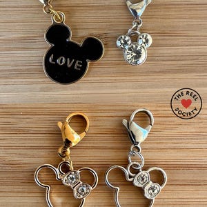 May include: Four charms with lobster clasps. Two charms are Mickey Mouse silhouettes, one black with "LOVE" and one with clear rhinestones. The other two are Minnie Mouse silhouettes, one gold and one silver, both with rhinestone bows.