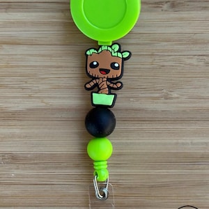 May include: A lime green retractable badge reel featuring a brown and green cartoon tree character charm. The design includes a matte black bead and a lime green bead below the character, finished with a clear plastic strap and a metal snap button.