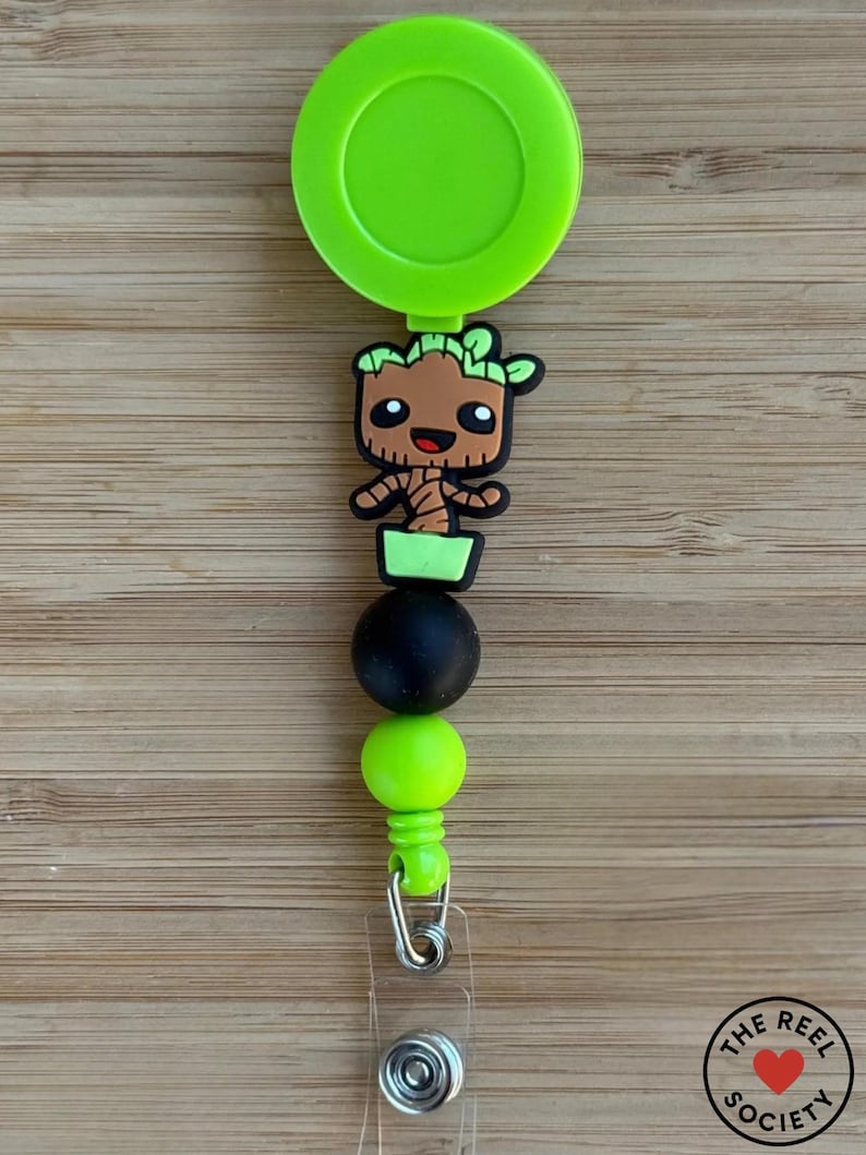 May include: A lime green retractable badge reel featuring a brown and green cartoon tree character charm. The design includes a matte black bead and a lime green bead below the character, finished with a clear plastic strap and a metal snap button.