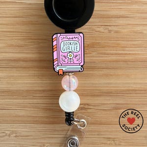 May include: A retractable badge reel with a black disc and a pink book-shaped charm that reads "Bookish Girlie." The charm is adorned with a flower and connected to a beaded strand with a white and pink bead. The Reel Society logo is visible.