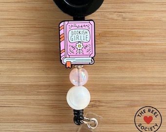 May include: A retractable badge reel with a black disc and a pink book-shaped charm that reads "Bookish Girlie." The charm is adorned with a flower and connected to a beaded strand with a white and pink bead. The Reel Society logo is visible.