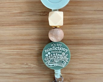 May include: A sage green retractable badge reel featuring a round topper with the white text it's too nonfiction outside and a graphic of an open book with flowers. The reel is accented with a cream geometric bead and a natural wood bead.