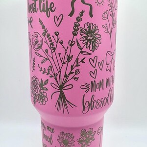 Peut inclure: Un gobelet rose avec un bord blanc, orn&eacute; de motifs floraux noirs, de n&oelig;uds, de c&oelig;urs et de textes tels que "Maman", "La vie est la meilleure vie" et "Amazing Mama". Le design enveloppe le gobelet isotherme.