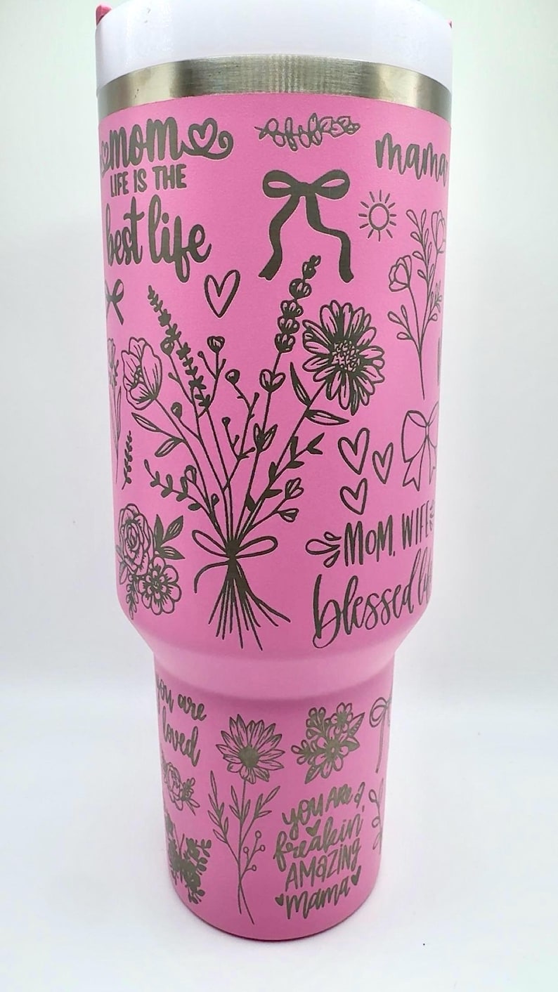 Peut inclure: Un gobelet rose avec un bord blanc, orn&eacute; de motifs floraux noirs, de n&oelig;uds, de c&oelig;urs et de textes tels que "Maman", "La vie est la meilleure vie" et "Amazing Mama". Le design enveloppe le gobelet isotherme.
