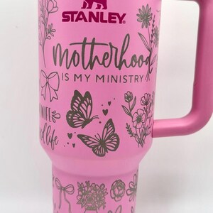 Peut inclure: Gobelet Stanley rose avec un couvercle et une poign&eacute;e blancs. Le gobelet est orn&eacute; de motifs floraux et de papillons, de c&oelig;urs et du texte "Motherhood is my ministry" et "mom". Le logo Stanley est visible.
