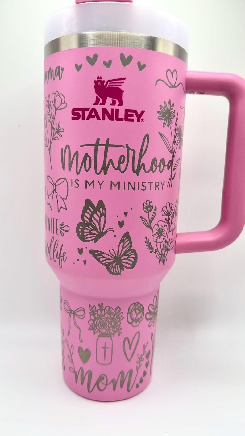 Peut inclure: Gobelet Stanley rose avec un couvercle et une poign&eacute;e blancs. Le gobelet est orn&eacute; de motifs floraux et de papillons, de c&oelig;urs et du texte "Motherhood is my ministry" et "mom". Le logo Stanley est visible.