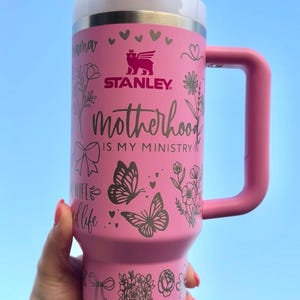 Peut inclure: Gobelet Stanley rose avec un couvercle blanc et une poign&eacute;e rose. Le gobelet porte l'inscription "Motherhood is my ministry" et des motifs d&eacute;coratifs floraux et papillons. Comprend une paille blanche.