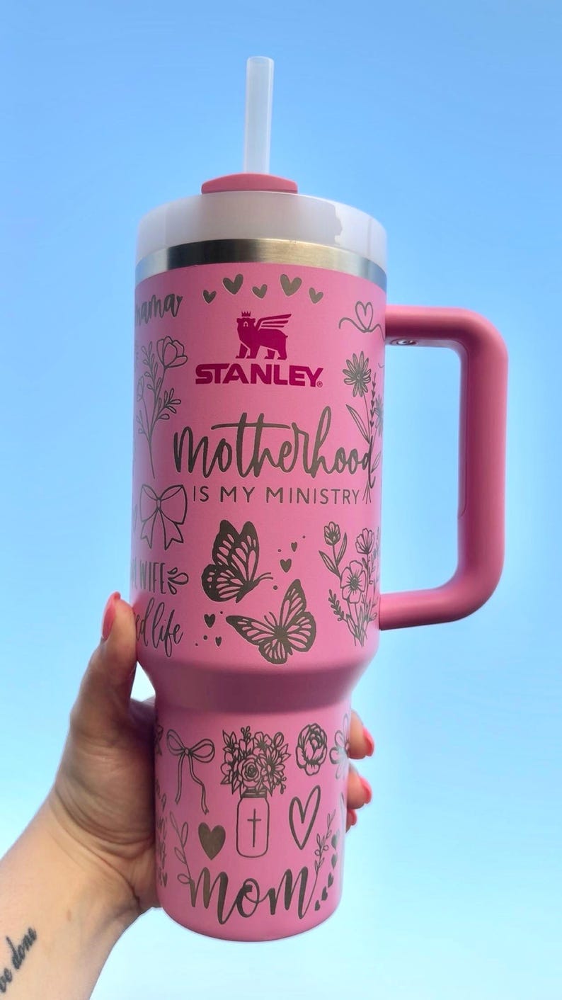 Peut inclure: Gobelet Stanley rose avec un couvercle blanc et une poign&eacute;e rose. Le gobelet porte l'inscription "Motherhood is my ministry" et des motifs d&eacute;coratifs floraux et papillons. Comprend une paille blanche.