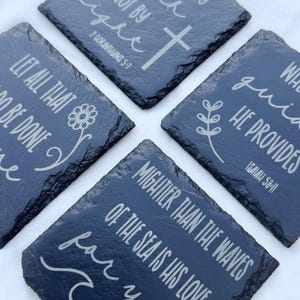 Puede incluir: Cuatro posavasos cuadrados de pizarra gris oscuro con citas inspiradoras y símbolos religiosos grabados en blanco. Cada posavasos presenta un versículo bíblico diferente, incluyendo una cruz, una flor y un diseño de olas.