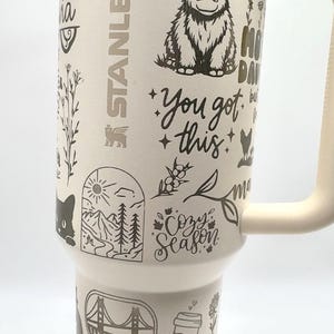 May include: Cream-colored insulated tumbler with a handle, featuring etched designs of a yak, the text "You got this," and other nature-themed illustrations. The tumbler also has the text "Cozy Season" and "Mama Bear."