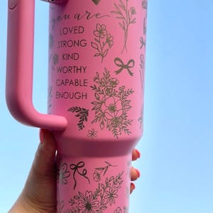 Peut inclure: Gobelet isotherme rose avec une poign&eacute;e, orn&eacute; de motifs floraux et des mots "you are loved, strong, kind, worthy, capable, enough". Le gobelet a un couvercle blanc et une poign&eacute;e rose.