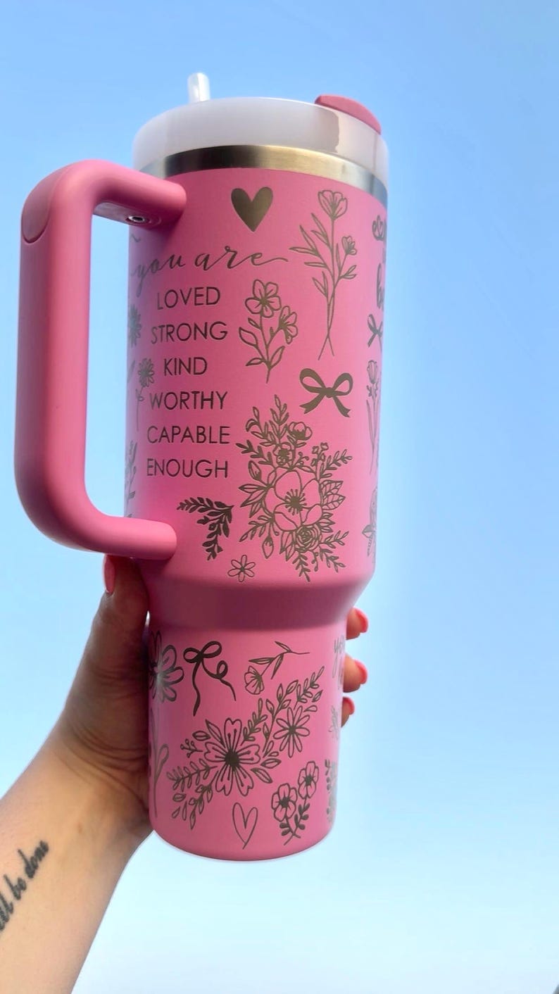Peut inclure: Gobelet isotherme rose avec une poign&eacute;e, orn&eacute; de motifs floraux et des mots "you are loved, strong, kind, worthy, capable, enough". Le gobelet a un couvercle blanc et une poign&eacute;e rose.
