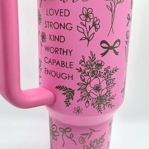 Peut inclure: Gobelet isotherme rose avec une poign&eacute;e, orn&eacute; de motifs floraux et de n&oelig;uds. Le gobelet porte les mots "you are LOVED STRONG KIND WORTHY CAPABLE ENOUGH" en &eacute;criture cursive. Comprend un symbole de c&oelig;ur.
